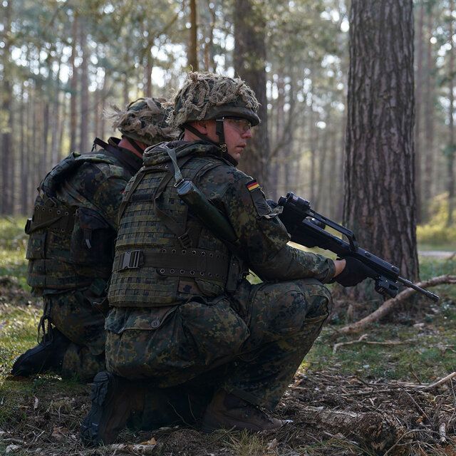 Zwei Soldaten in Tarnkleidung und mit Gewehr befinden sich in einem Wald