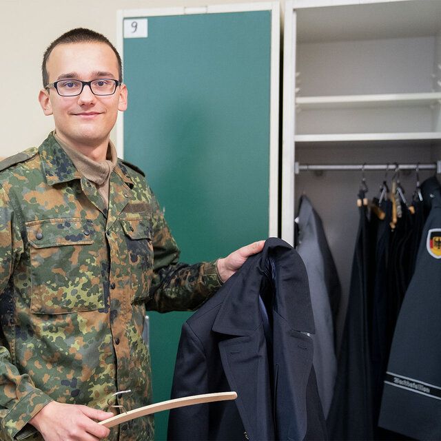 Ein Soldat in Tarnkleidung steht vor einem Kleiderschrank und hält eine Jacke und einen Kleiderbügel in der Hand