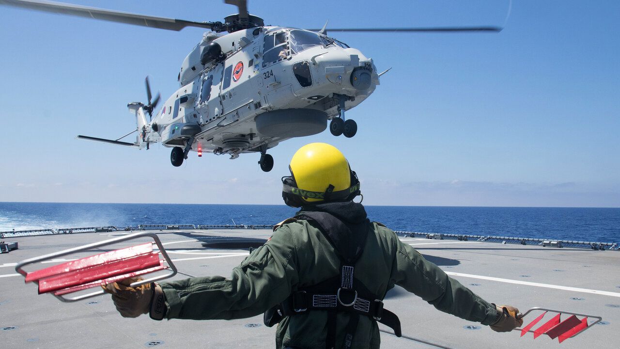 Ein Hubschrauber der Marine landet auf einem Schiff. Ein Soldat unterstÃ¼tzt ihn und weist ihn ein.