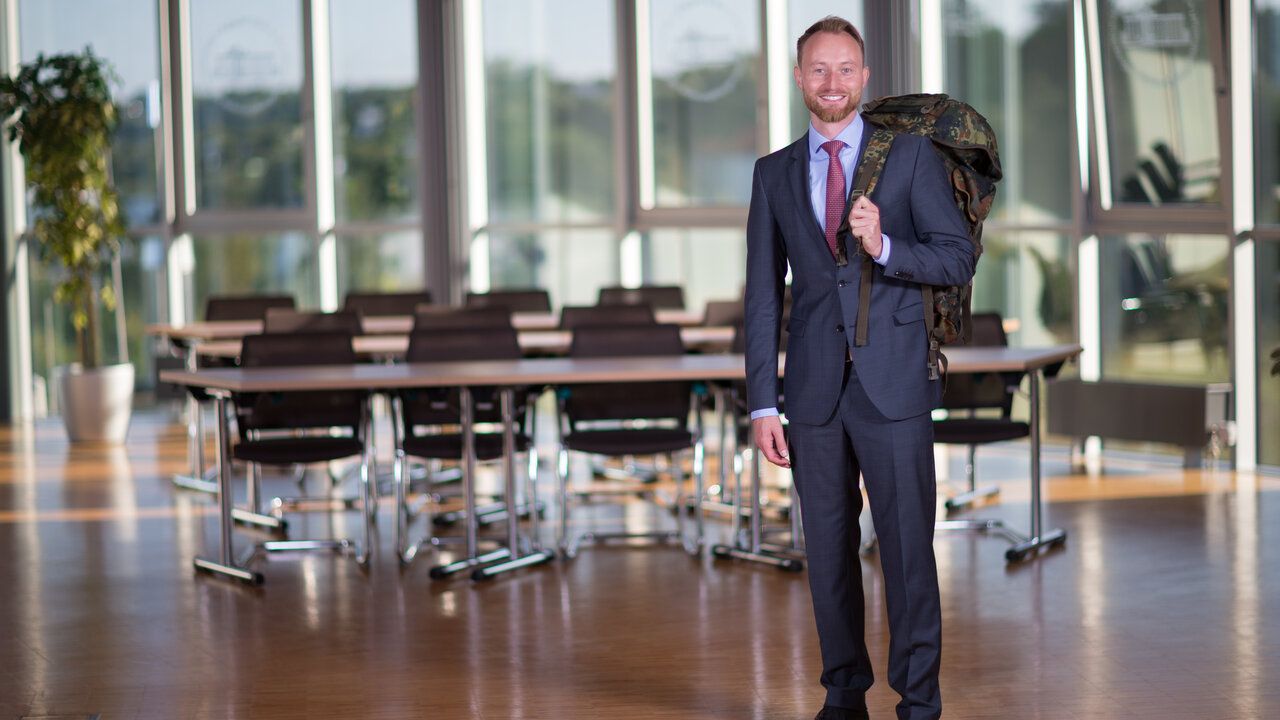Ein Mann im Anzug und einem Rucksack der Bundeswehr steht in einem Saal. 