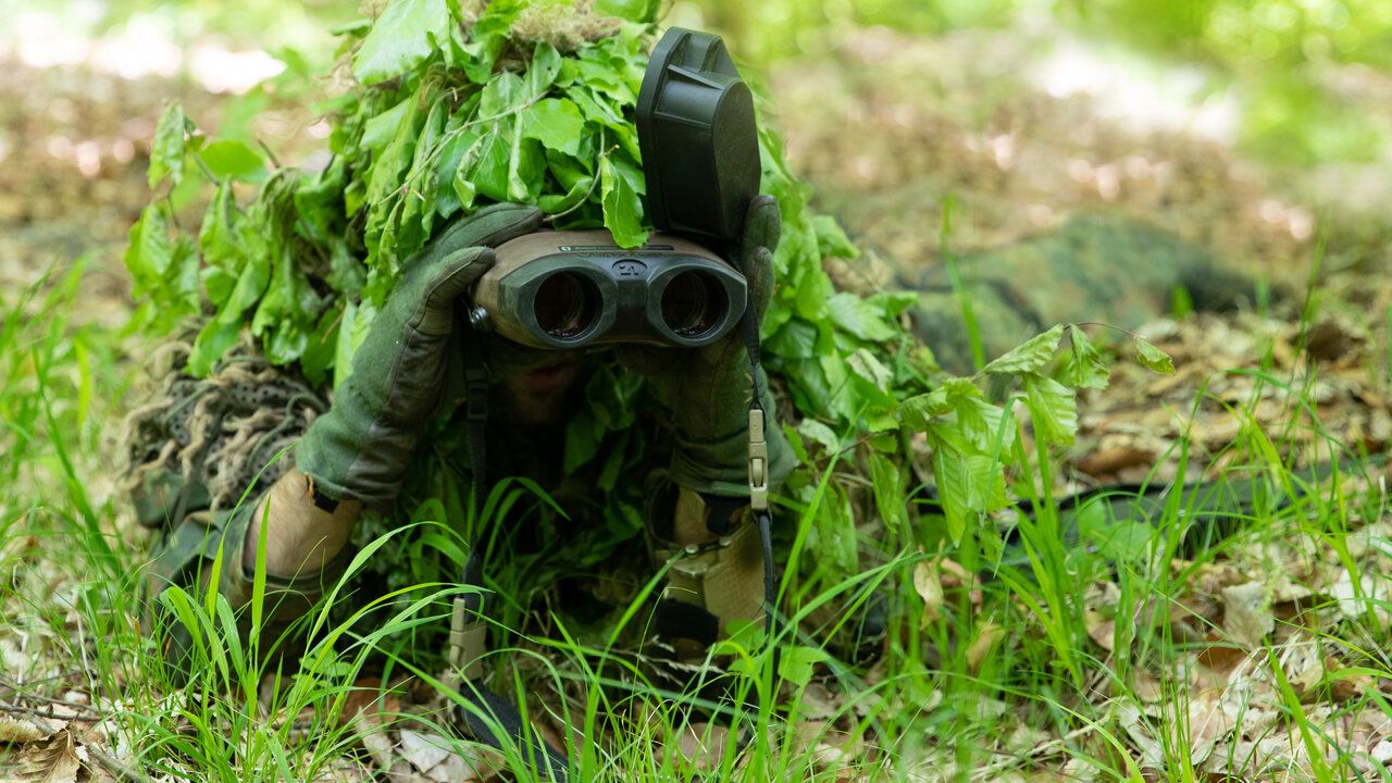 Ein Soldat auf dem Waldboden beobachtet mit einem Fernglas das Geschehen. Er ist durch grÃ¼ne BlÃ¤tter so getarnt das man ihn schwer erkennen kann.