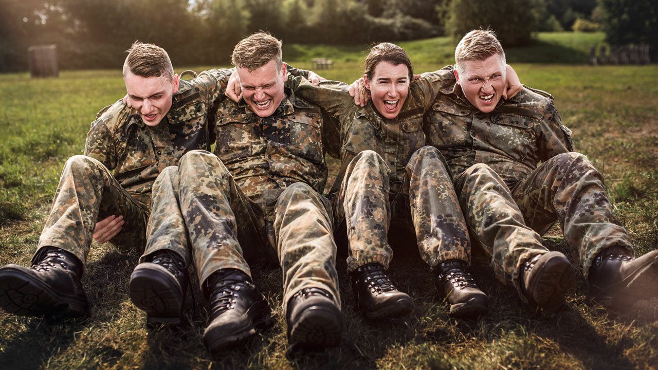 Soldat_innen in Uniform sitzen Arm in Arm auf dem Boden und helfen sich gegenseitig sich hochzuziehen 