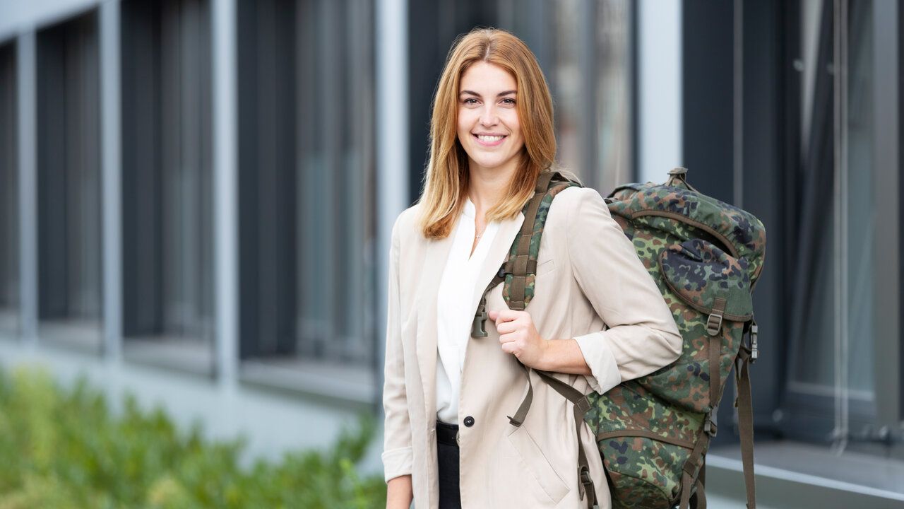 Eine Frau in zivil Kleidung steht mit einem Bundeswehrrucksack vor einem Gebäude