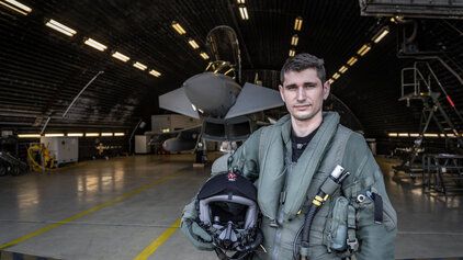 Pilot der Bundeswehr steht mit Helm unter dem Arm im Hangar, im Hintergrund ist ein Jet zu sehen.