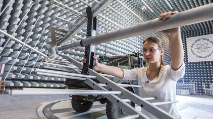 Eine Frau im Vordergrund baut eine Metallkonstruktion fÃ¼r einen Versuch auf. Sie ist in einer Halle, in welcher man elektromagnetische VertrÃ¤glichkeiten messen kann. Die WÃ¤nde sehen aus wie die OberflÃ¤chen eines Eierkartons