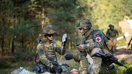 Eine Soldatin und ein Soldat versorgen einen verwundeten Kameraden an einem WaldstÃ¼ck. 