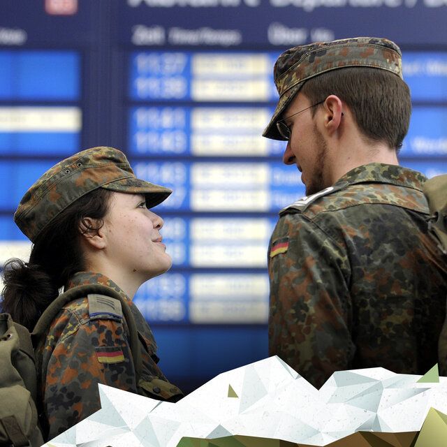 Eine Soldatin und ein Soldat stehen vor einer Anzeigetafel der deutschen Bahn. 