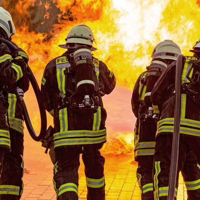4 Feuerwehrleute stehen vor einem Feuer und versuchen dieses mit ihren SchlÃ¤uchen zu lÃ¶schen. 