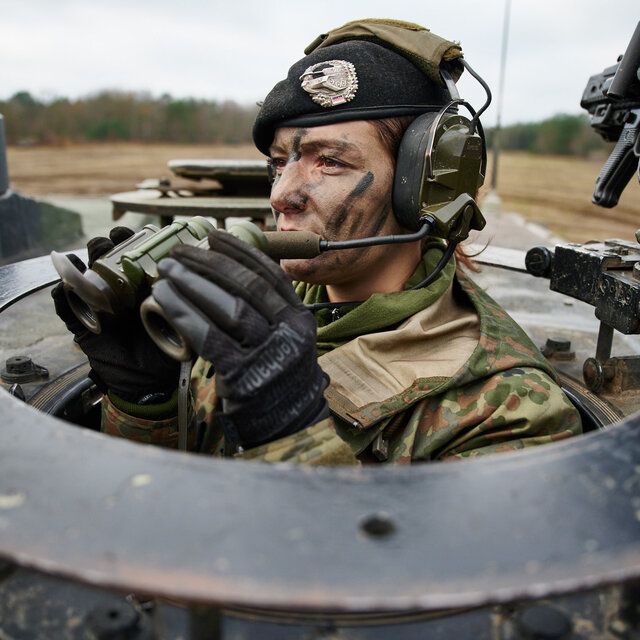 Eine Soldatin beobachtet mit einem Fernglas die Lage aus einem Panzer. Sie trÃ¤gt Tarnschminke und ihr Barett.