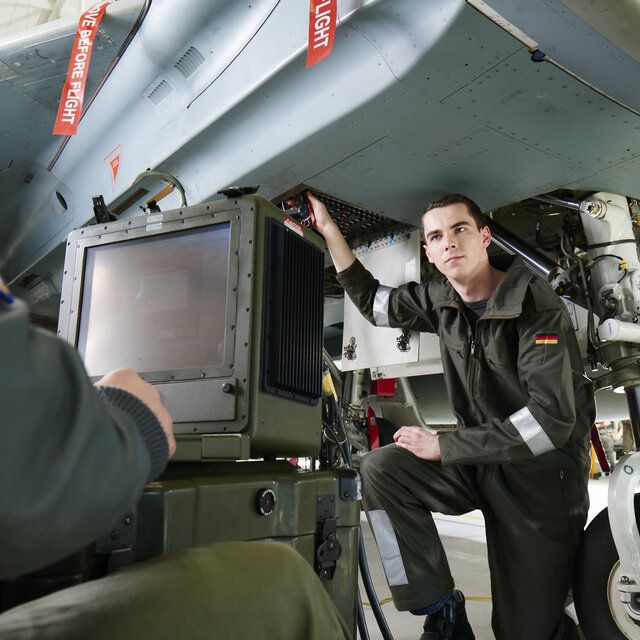 Ein FluggerÃ¤tmechaniker und ein Soldat arbeiten unter einem Eurofighter. Der Soldat sitzt dabei vor einem Monitor wÃ¤hrend der FluggerÃ¤tmechaniker die FunktionalitÃ¤t prÃ¼ft. 