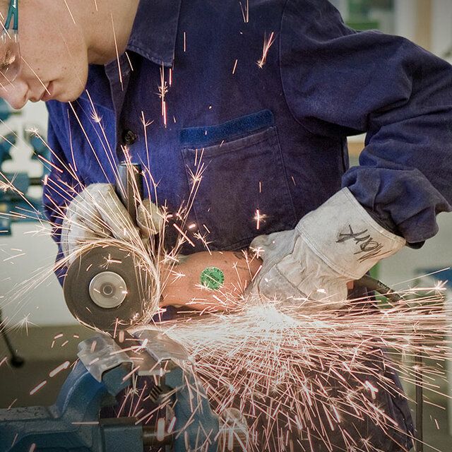 Ein Mechaniker schleift in einer Werkstatt Metall. Funken treten auf doch durch seine Arbeitskleidung und Schutzbrille ist er gut geschÃ¼tzt. 