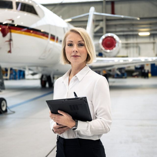 Eine Ingenieurin steht in einer Halle vor einem Flugzeug. In ihren Händen trägt sie eine schwarze Mappe. 