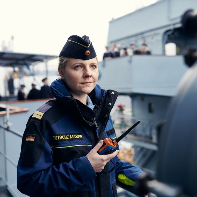 Eine Offizierin der Marine steht mit einem FunkgerÃ¤t vor einem Schiff. 