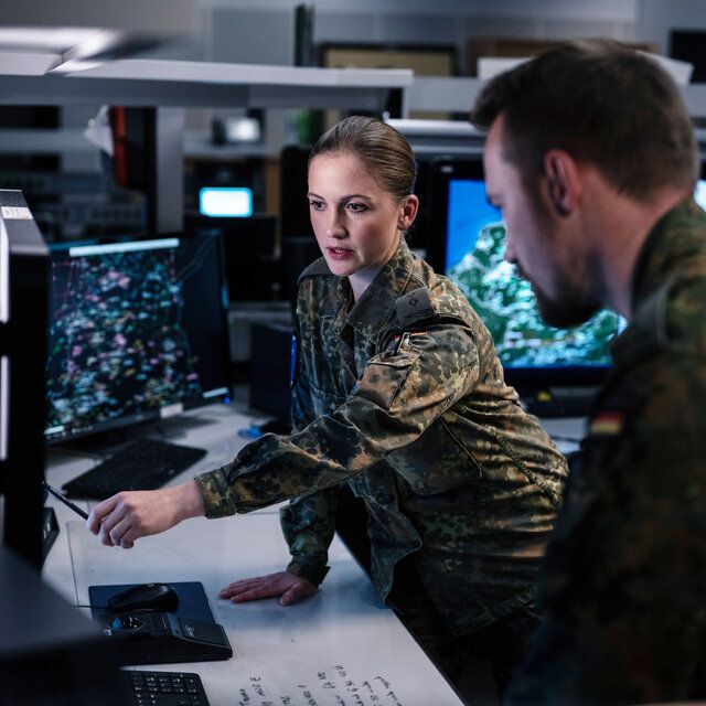 Eine Soldatin und ein Soldat der Bundeswehr besprechen sich vor einem Monitor. Die Soldatin zeigt mit ihrer Hand auf einen der Monitore.