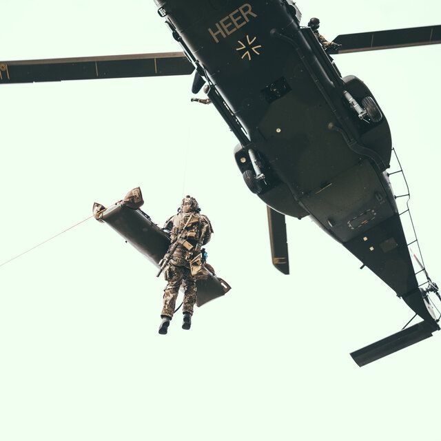 Ein verwunderter Soldat_in wird mit dem Hubschrauber gerettet. Ein Soldat_in seilt sich dazu mit einer Trage aus dem Hubschrauber ab.