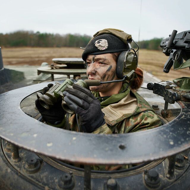 Eine Soldatin im Panzer schaut mit einem Fernglas aus der Lucke geraus.